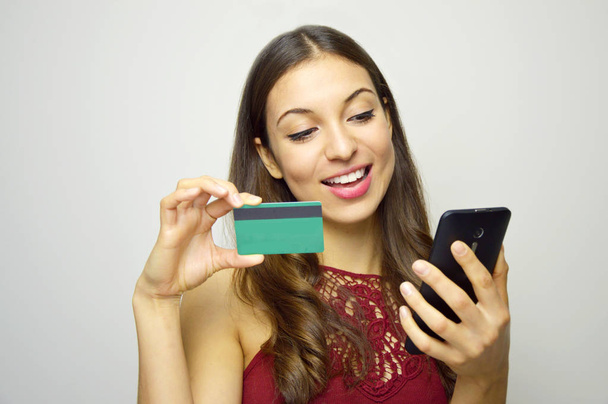glücklich lächelndes Mädchen, das sein Smartphone mit Kreditkarte in der anderen Hand auf weißem Hintergrund hält und zu ihr blickt. E-Commerce-Frau. Menschen, die online einkaufen. - Foto, Bild
