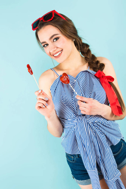 Happy pretty brunette girl holding two heart shaped lollypops on blue studio background  - Fotografie, Obrázek