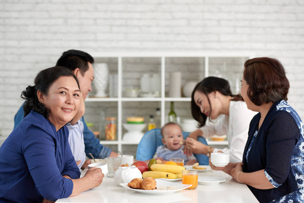 Портрет зрелой азиатки, сидящей за обеденным столом с большой семьей и улыбающейся в камеру
 - Фото, изображение