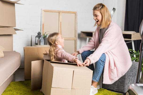 happy mother and daughter looking at each other while playing with cardboard boxes during relocation - Photo, Image