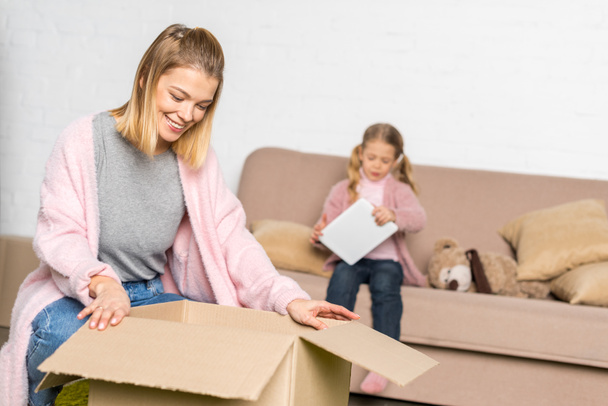 Lachende vrouw verpakking kartonnen doos terwijl dochtertje met behulp van digitale tablet - Foto, afbeelding