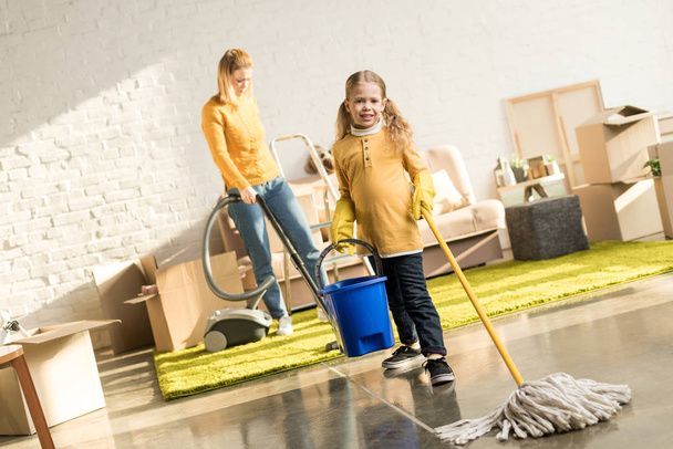 mother and daughter with mop and vacuum cleaner cleaning room while moving home - Photo, Image