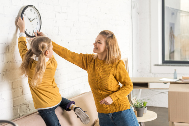 happy mother and daughter hanging clock on wall during relocation    - Foto, afbeelding