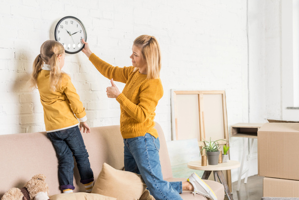 Mutter und Tochter hängen beim Umzug Uhr an Wand  - Foto, Bild