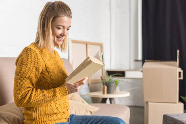 smiling young woman reading book while moving home - Foto, afbeelding