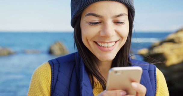 Close up of beautiful young woman texting on the smart phone on and smiling the rocky sea background. Cold sunny weather. Outdoors. Portrait shot - Foto, Imagem