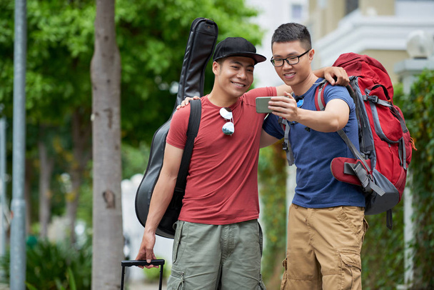 Handsome young tourists with wide smiles taking selfie on smartphone while walking in picturesque town - Photo, Image
