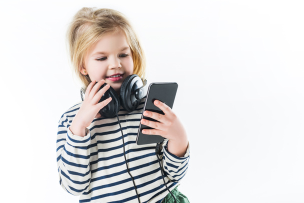 χαμογελαστό μικρό παιδί με τα ακουστικά και το smartphone που απομονώνονται σε λευκό - Φωτογραφία, εικόνα