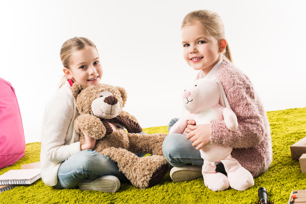 hermanas pequeñas felices jugando con juguetes suaves juntas mientras están sentadas en el piso aisladas en blanco
 - Foto, Imagen