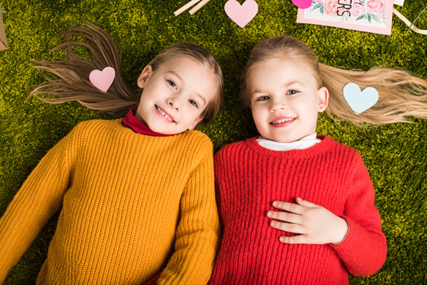 Draufsicht auf glückliche kleine Schwestern, die auf Teppich liegen, umgeben von Herzen - Foto, Bild