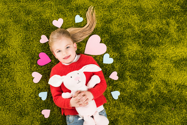 top view of adorable little child surrounded with hearts lying on green soft carpet with toy rabbit - Photo, Image