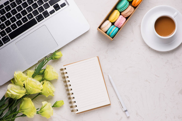 Escritorio blanco con macarrones de colores, flores, cuaderno, portátil y taza de café
 - Foto, Imagen
