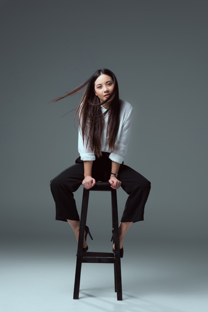 beautiful young asian woman sitting on chair and looking at camera on grey - 写真・画像