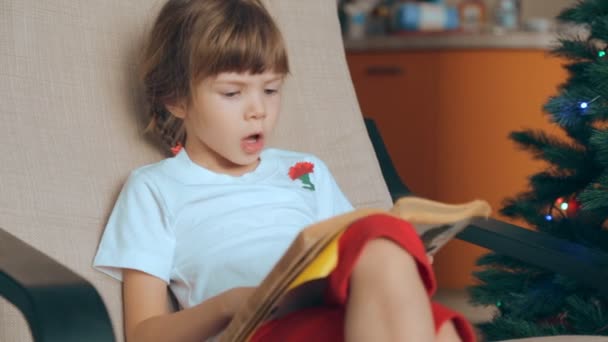 Small girl reading a book in front of Christmas tree - Záběry, video