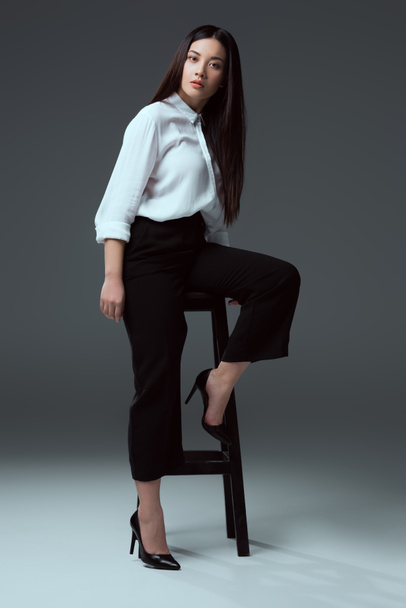 full length view of beautiful stylish asian girl posing with chair and looking at camera on grey - Foto, immagini