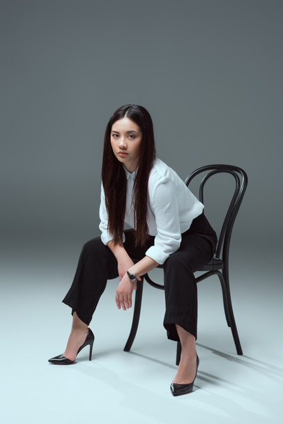full length view of attractive asian girl sitting on chair and looking at camera on grey  - Fotografie, Obrázek