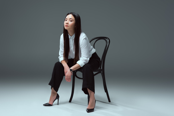 full length view of beautiful young asian woman sitting on chair and looking at camera on grey  - Фото, зображення