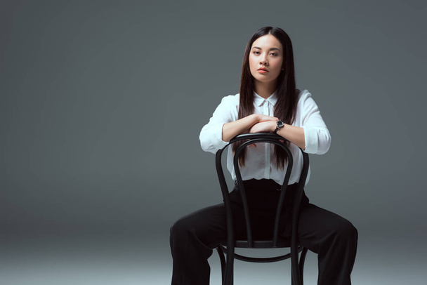 beautiful asian girl sitting on chair and looking at camera on grey - Φωτογραφία, εικόνα