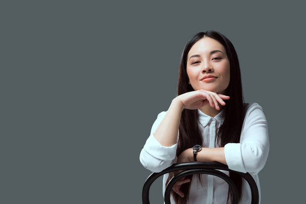 beautiful asian girl sitting on chair and smiling at camera isolated on grey  - Zdjęcie, obraz