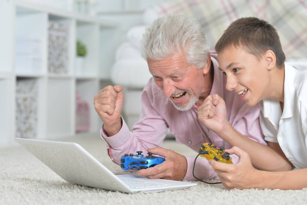 Дедушка и внук лежат на полу и играют в компьютерные игры на ноутбуке
 - Фото, изображение