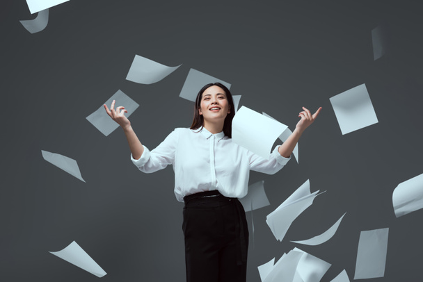 glimlachend jonge zakenvrouw gooien papieren geïsoleerd op grijs - Foto, afbeelding