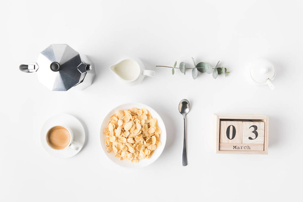 acostado plano con hojuelas de maíz en un tazón para el desayuno, cafetera y taza de café en la superficie blanca
 - Foto, Imagen