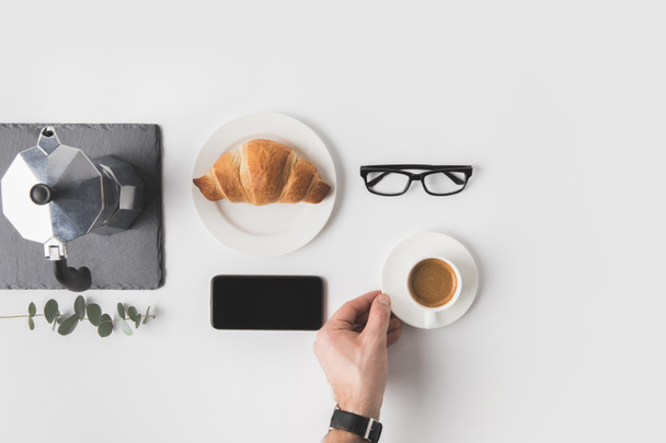cropped shot of man holding cup of coffee with smartphone, eyeglasses and croissant for breakfast near by on white surface - Foto, afbeelding