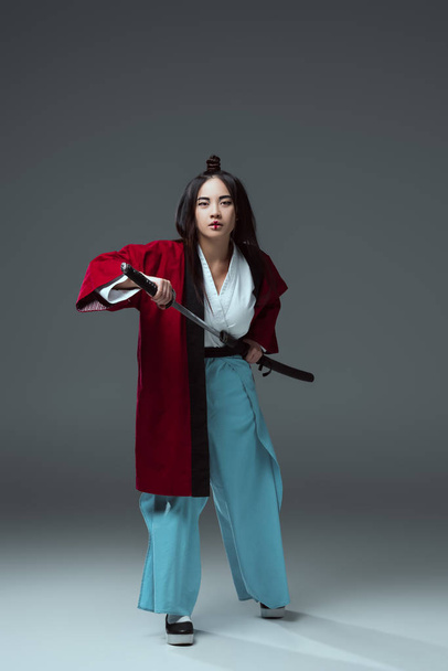 full length view of young asian woman in kimono holding katana sword and looking at camera on grey - Foto, Imagem