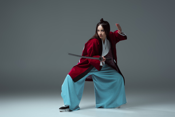 young asian woman in kimono fighting with katana sword and looking away on grey - Zdjęcie, obraz