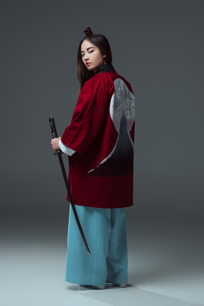 back view of woman in traditional japanese kimono holding katana and looking at camera on grey   - Photo, Image