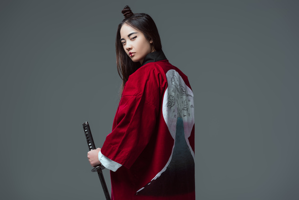 young woman in traditional japanese kimono holding katana and looking at camera isolated on grey  - Fotografie, Obrázek