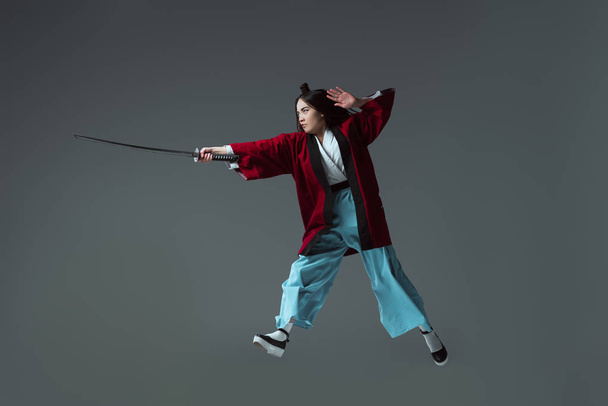 full length view of samurai in kimono fighting with katana in jump isolated on grey - 写真・画像