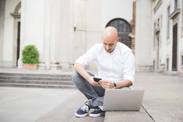 Jeune homme d'affaires contemporain travaillant à distance assis à l'extérieur dans la ville en utilisant un smartphone et un ordinateur
 - Photo, image