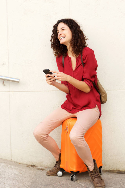Teljes test portré fiatal nő ül a bőröndöt, és a gazdaság smartphone - Fotó, kép