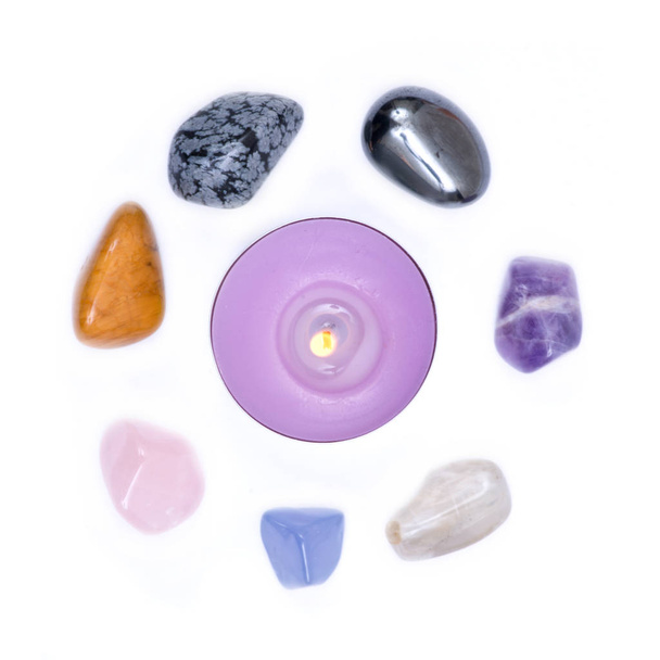 Лавандовая чайная свеча окружена набором из семи целебных камней чакры для кристального заживления, изолированных на белом фоне
  - Фото, изображение