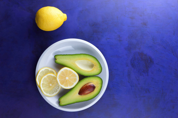 Avocado and slices of lemon on a white plate, tropical fruits on an ultraviolet background, vegetarian breakfast, Asian cuisine, avocado pop art, citrus with avocado, art - Fotografie, Obrázek