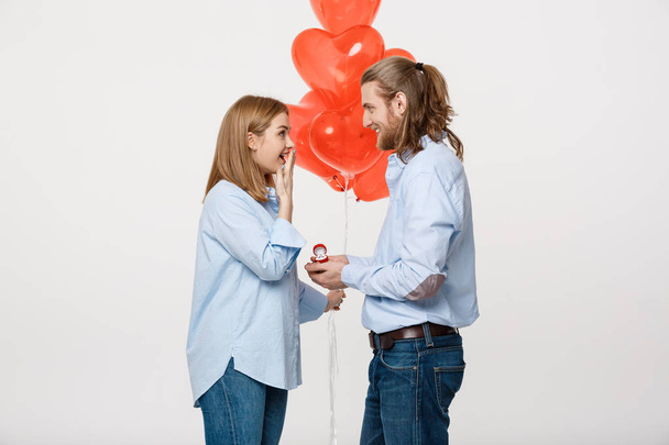 Retrato de joven chico guapo da un anillo a una chica sobre un fondo blanco con globos de aire rojo corazón
. - Foto, Imagen