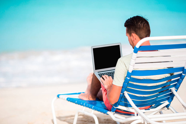 Young man with laptop on tropical caribbean beach. Man sitting on the sunbed with computer and working on the beach - Photo, Image