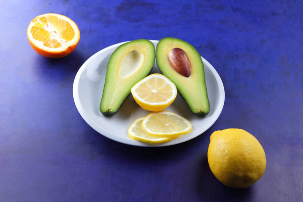 Avocado, lemon and half orange on a white plate, tropical fruits on an ultraviolet background, vegetarian breakfast, Asian cuisine, avocado pop art, citrus, orange with avocado, art - Фото, изображение