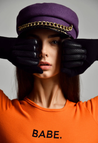 Portrait of young sexy brunette woman in purple peaked cap or beret with gold chain in leather gloves and orange shirt - Фото, изображение