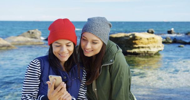 Pretty young women making nice selfies on the smart phone and watching them then. The rocks and sea background. Cold but sunny day. Outdoor - Photo, image