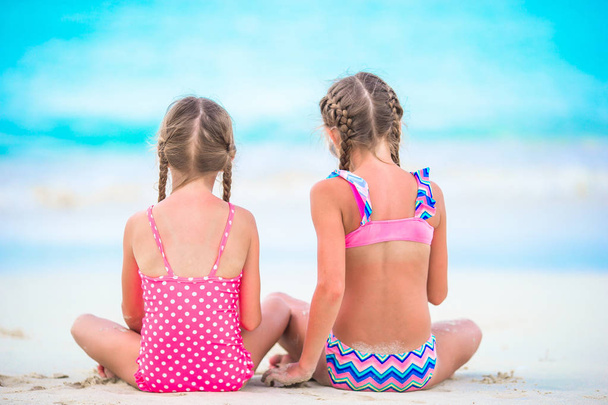 砂浜で砂遊びをする愛らしい少女たち。浅い水の中に座って砂の城を作る子供たちのバックビュー - 写真・画像