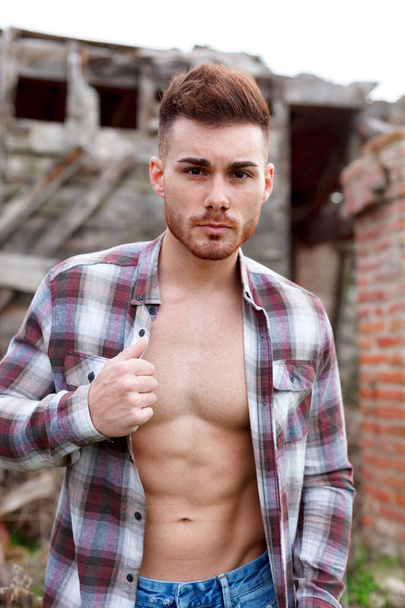 attractive guy wearing plaid shirt unbuttoned against abandoned house - Photo, Image