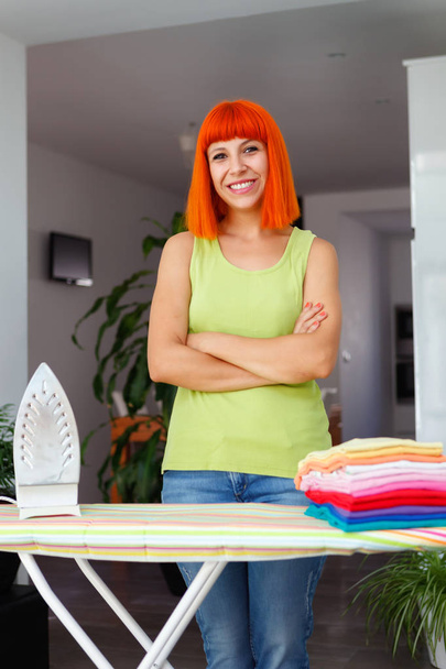 Рыжая красивая молодая женщина улыбается и гладит одежду дома
 - Фото, изображение