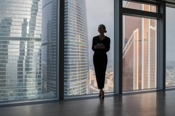 Zakenvrouw holding van een tablet en permanent in een modern kantoor. Panoramische ramen achtergrond. - Foto, afbeelding