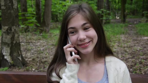 Young attractive girl speak to phone on bench. Summer park. Smile. HD video footage shooting static camera. - Materiał filmowy, wideo
