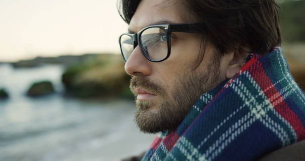 Close up of a appealing sad man in glasses thinking on the rocky beach. Cloudy cold day. Outdoors - Photo, Image