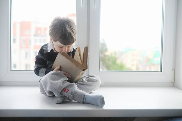 O rapazinho está a ler um livro. A criança senta-se na janela de um
 - Foto, Imagem