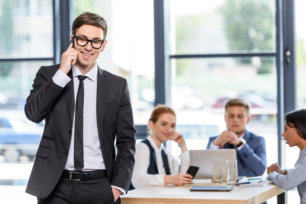 Hombre de negocios sonriente hablando en el teléfono inteligente delante de sus colegas en la oficina moderna
 - Foto, Imagen