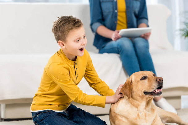 excited little kid petting dog on floor while mother using tablet on couch - Photo, Image